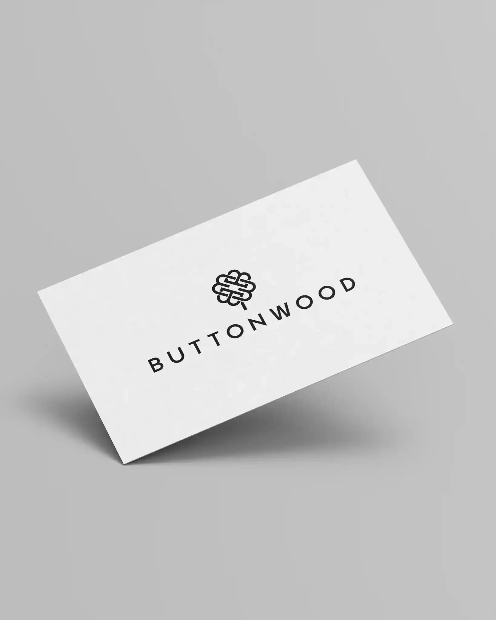 Close-up of a white card with the word "BUTTONWOOD" and a logo with a knot design printed on it, placed on a grey surface.