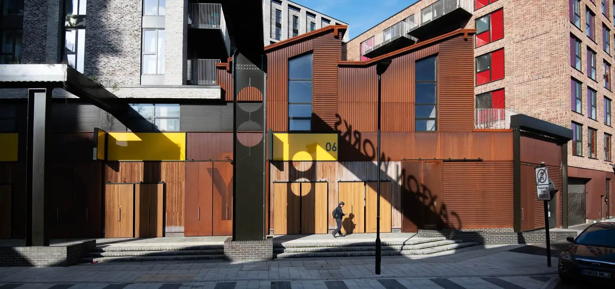 Modern multi-story building with a mix of brick and wood exterior, featuring large windows, balconies, and brightly colored panels. Pedestrian crossing sign and a person walking on the sidewalk.