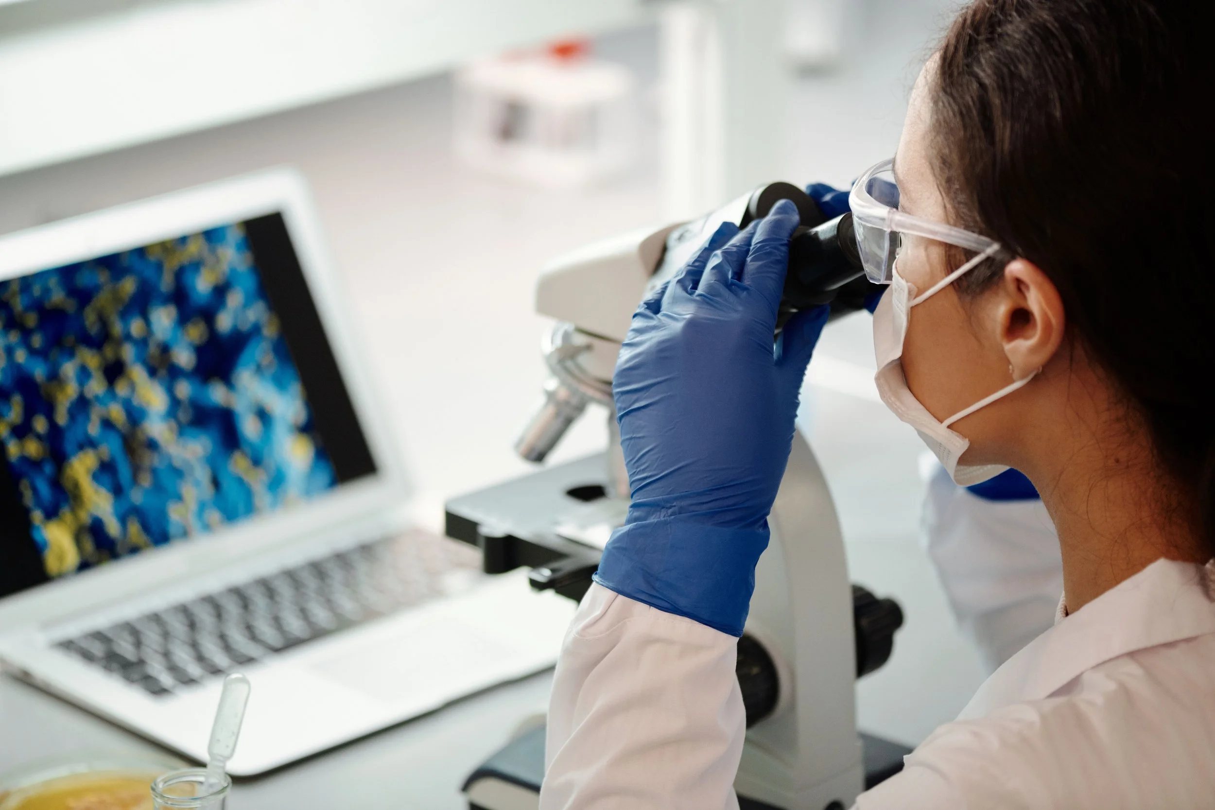 A female scientist wearing safety goggles, gloves, and a face mask looking into a laboratory microscope.