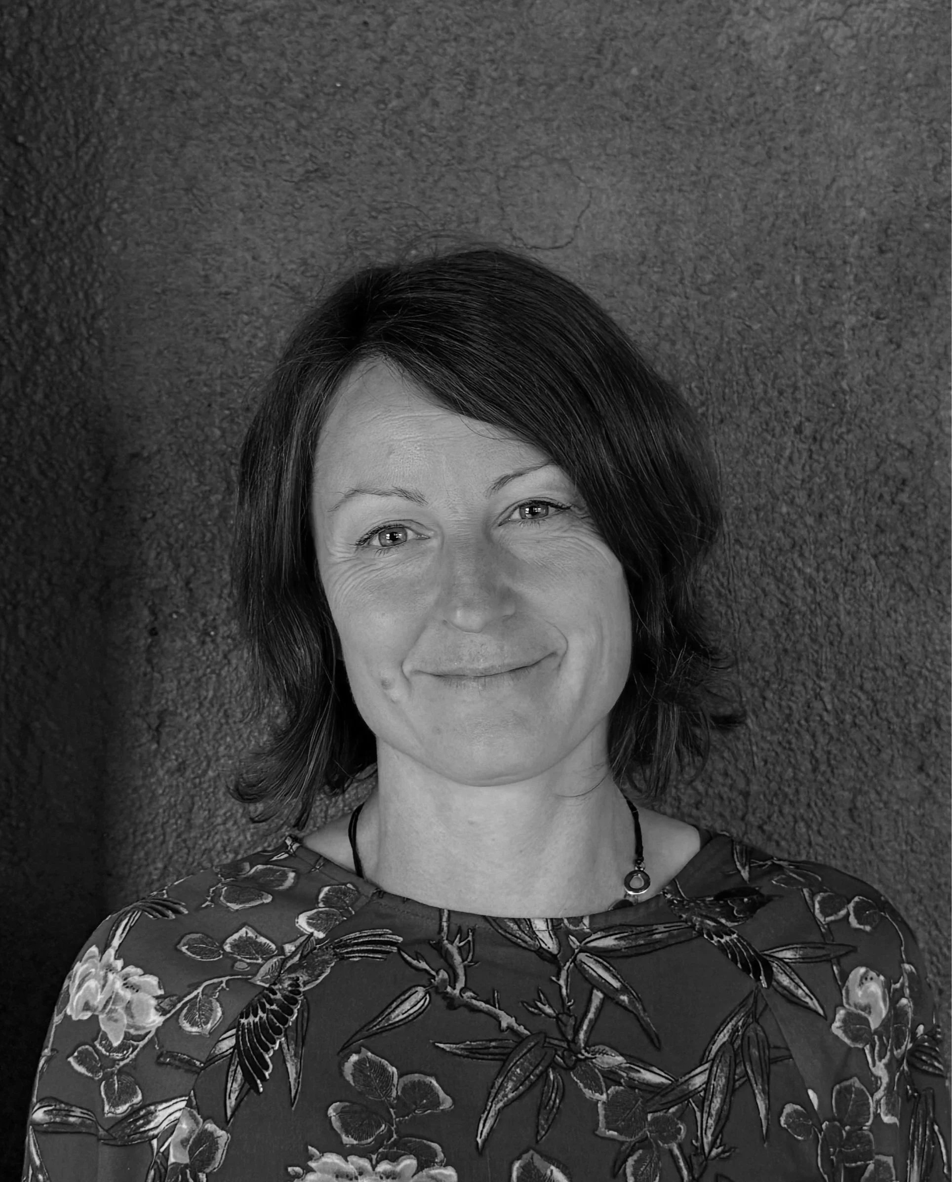 A black and white photo of a smiling woman with short dark hair, wearing a patterned top and a necklace, lying on a textured surface.