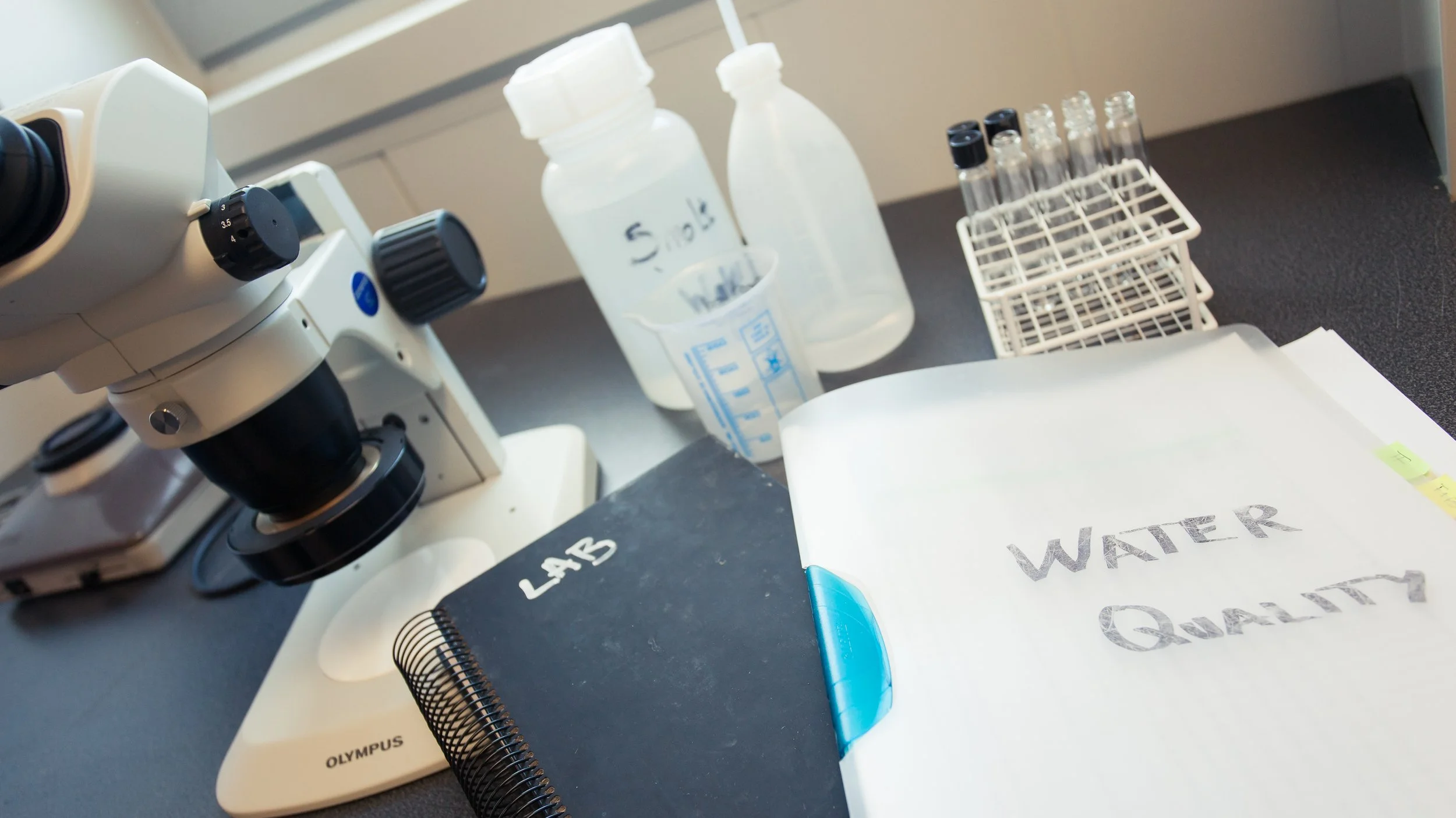 A laboratory workspace with a microscope, bottles of water, a test tube rack, labeled papers, and a blue pen.