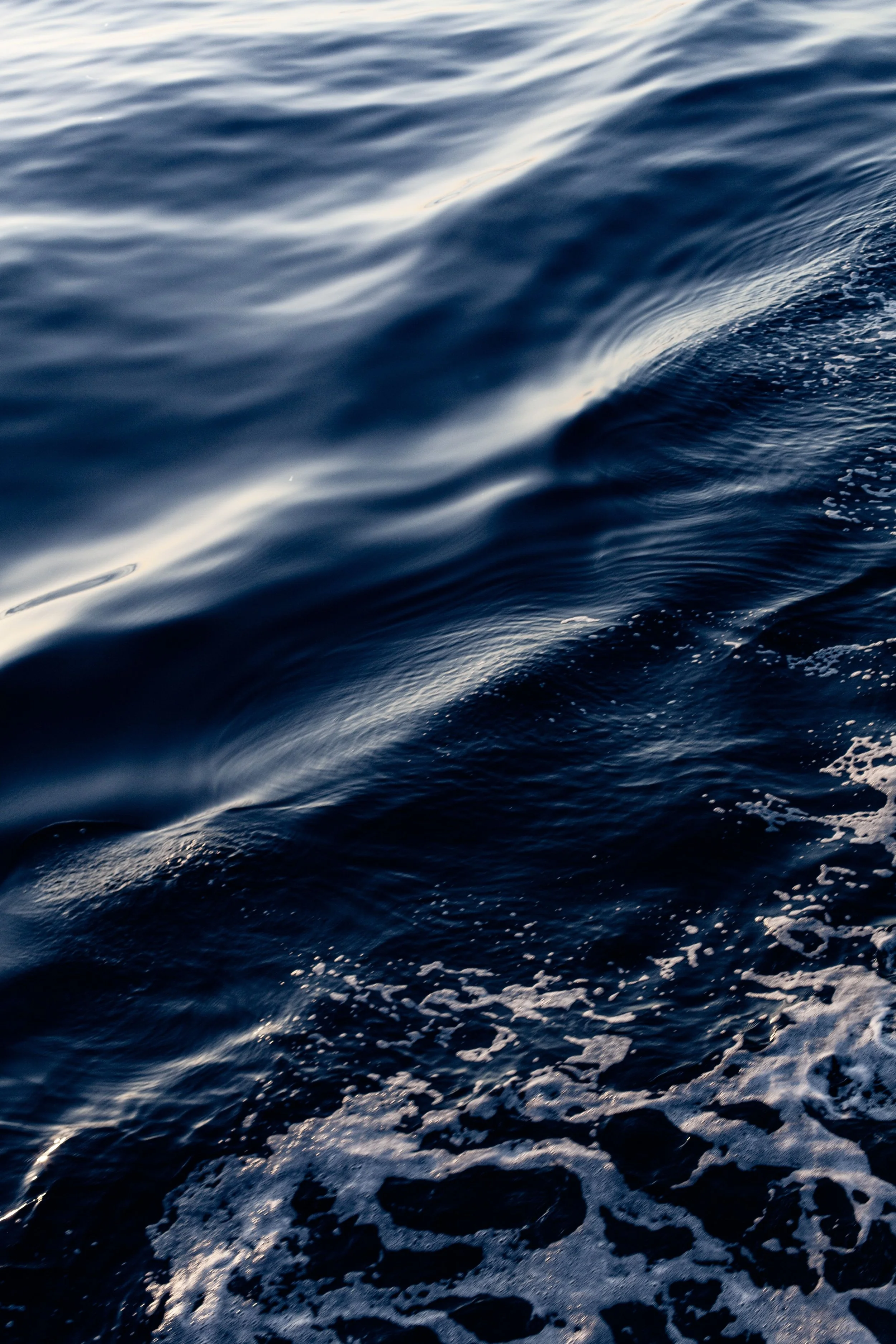 Close-up of ocean waves with white foam on the surf and dark blue water under bright light.