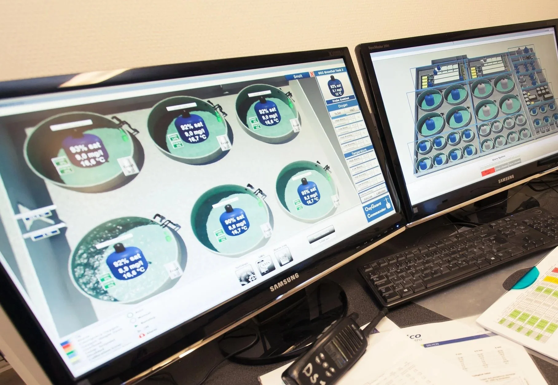 Two computer monitors displaying an industrial control system screen for monitoring oxygen tanks, with a keyboard, remote control, and printed documents on a desk.