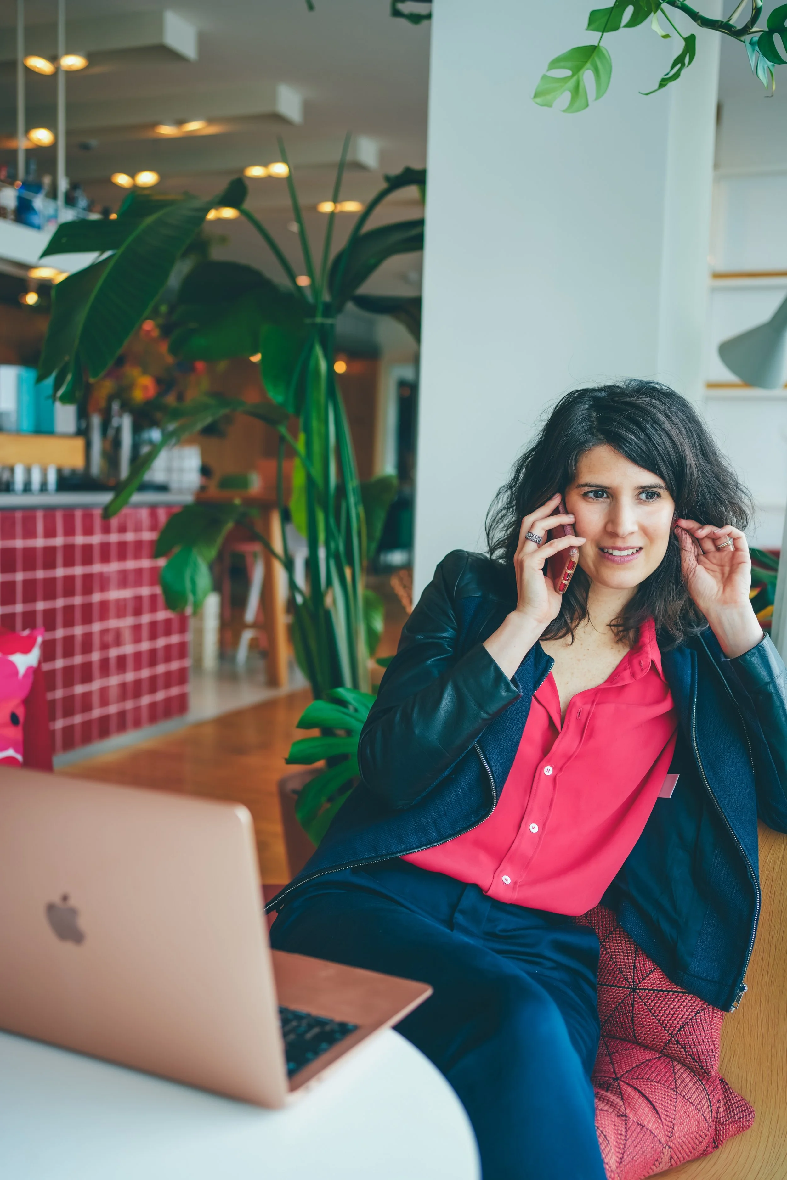 Une femme parlant au téléphone dans un café, assise à une table avec un ordinateur portable Apple. Elle porte une veste en cuir et une chemise rose. La scène est ornée de plantes vertes et d'un décor confortable.