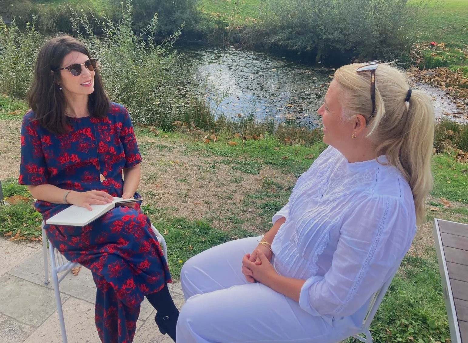 Deux femmes discutant assises à côté d'un petit étang en plein air, avec des arbres en arrière-plan, l'une portant une robe rouge et bleu, l'autre une chemise blanche, portant des lunettes.