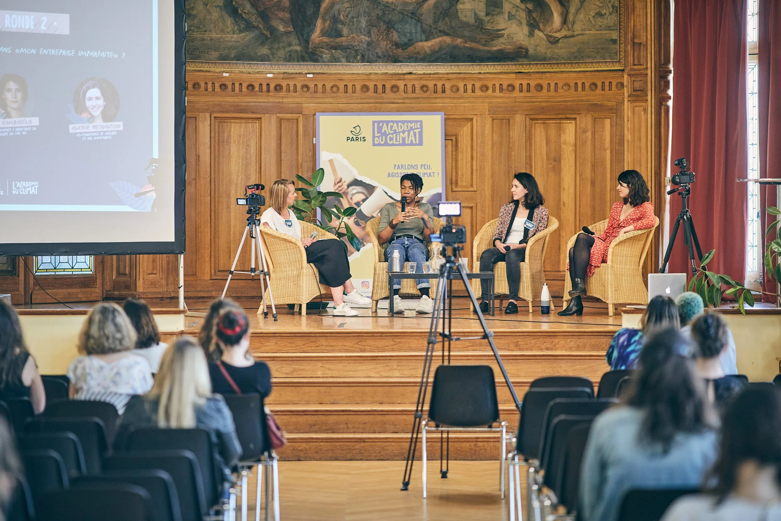 Une scène d'une conférence ou table ronde dans une salle avec un décor en bois, quatre femmes assises en discussion, des caméras et des microphones, et un public attentif. Il y a un panneau indiquant 'L'Académie du Climat'.
