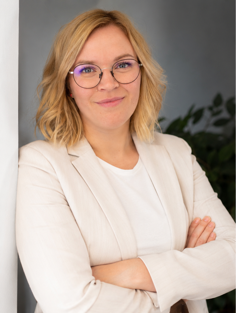 Frau mit Brille, blondes Haar, weißen Blazer, lächelnd, im Büro mit Pflanzen im Hintergrund.