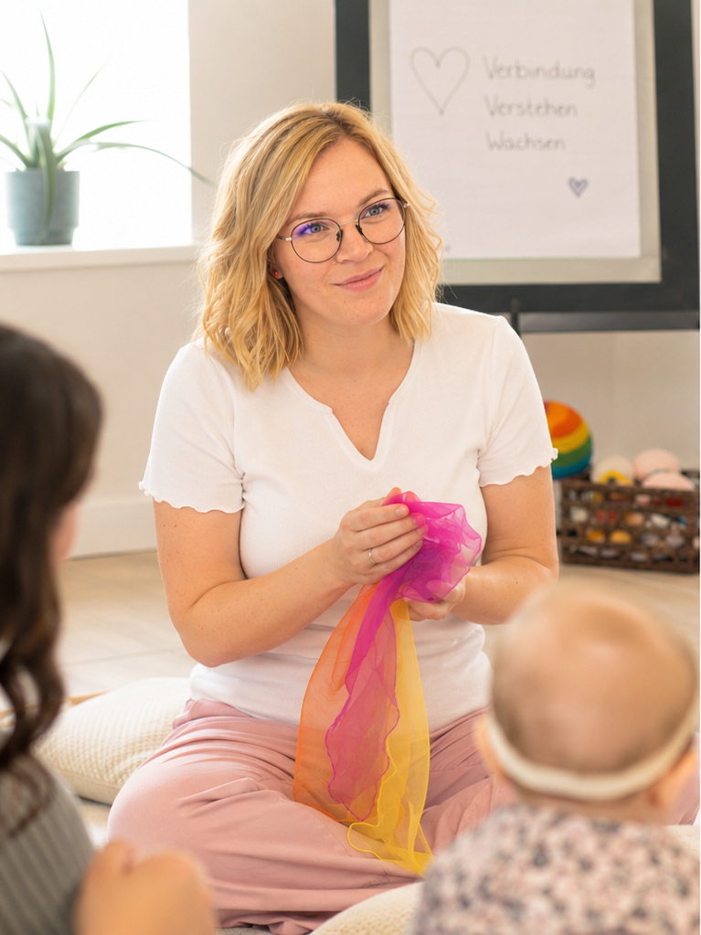 Eine Frau mit Brille und blonden Haaren sitzt auf dem Boden, hält bunte Stoffstücke in den Händen und spricht mit zwei Kindern. Im Hintergrund ist eine Wand mit einem Whiteboard, auf dem Herz und Wörter geschrieben sind.