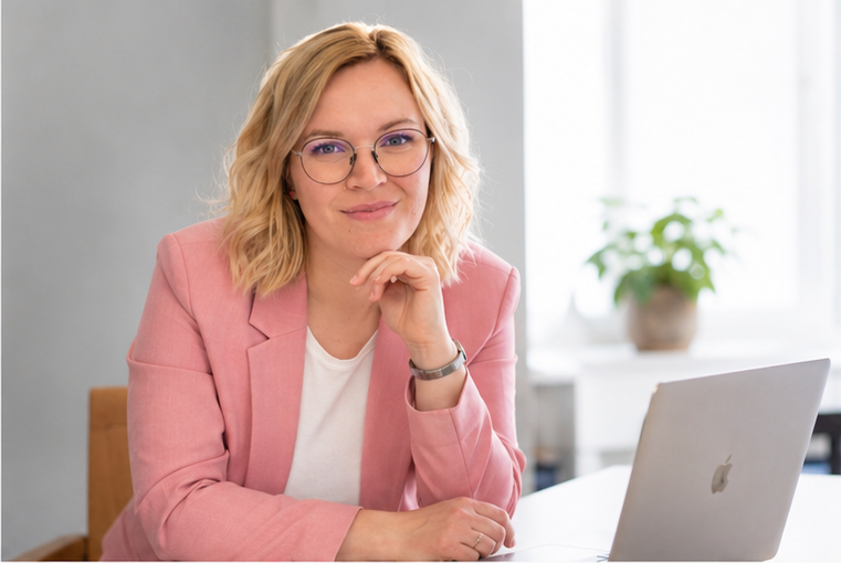 Eine Frau mit Brille, blondem Haar und pinkem blazer sitzt an einem Schreibtisch mit einem Laptop und lächelt.