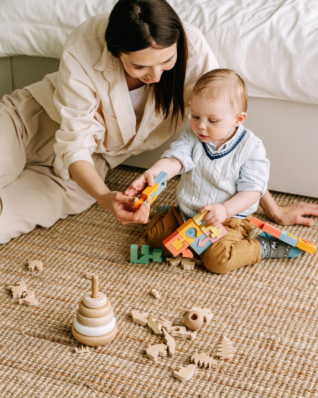 Eine Frau spielt mit einem kleinen Jungen, der bunte Holzbausteine hält, auf einem Teppich. Es gibt eine Holz-Wiege und andere Spielsachen um sie herum.