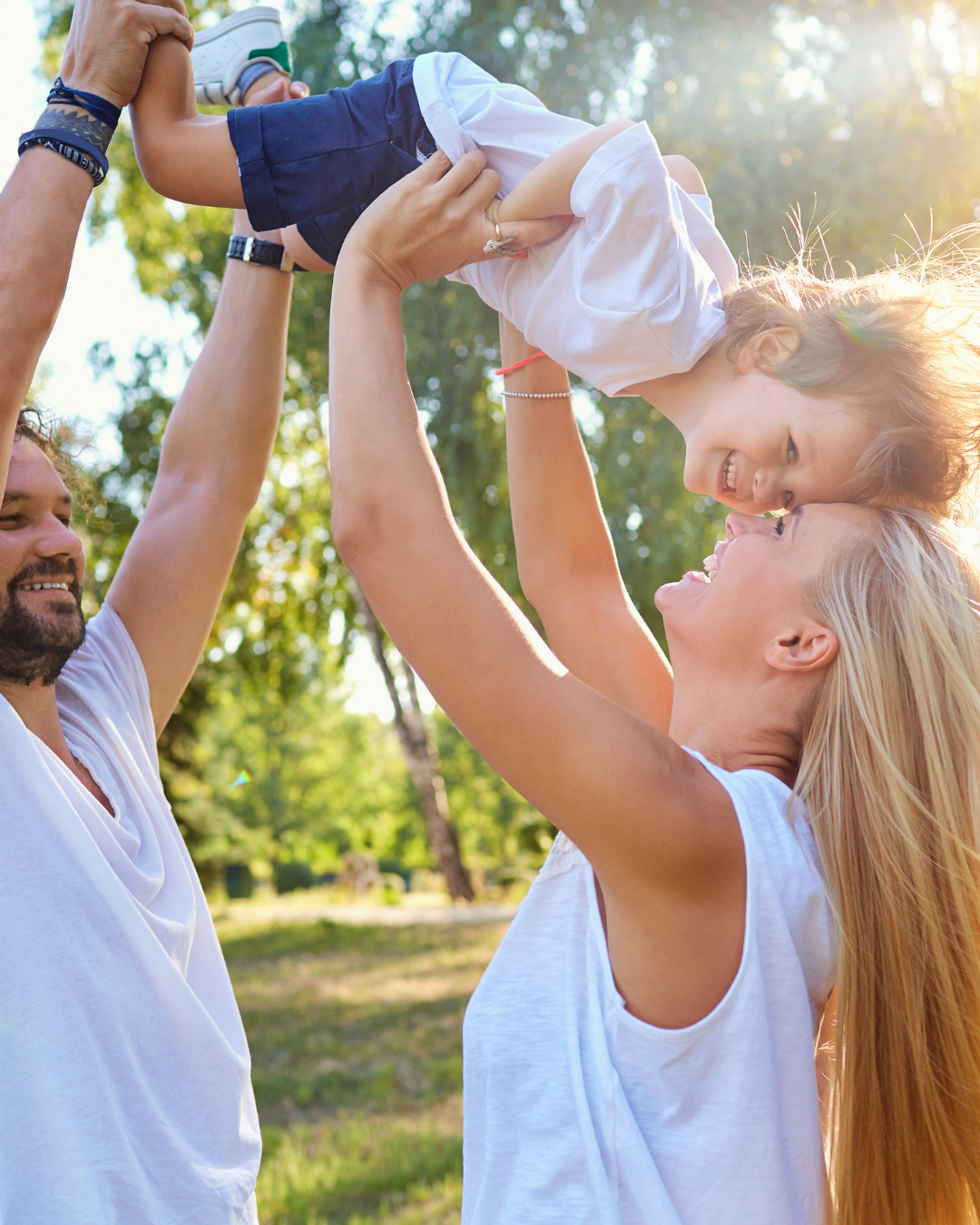 Familie beim Spielen im Park, Eltern heben Kind in die Luft, Sonne scheint durch Bäume.