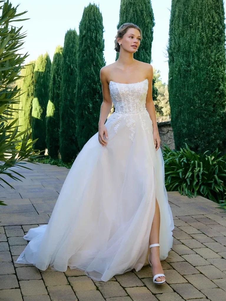 A woman in a strapless white wedding gown with lace details, standing outdoors on a brick pathway with tall cypress trees and greenery in the background.