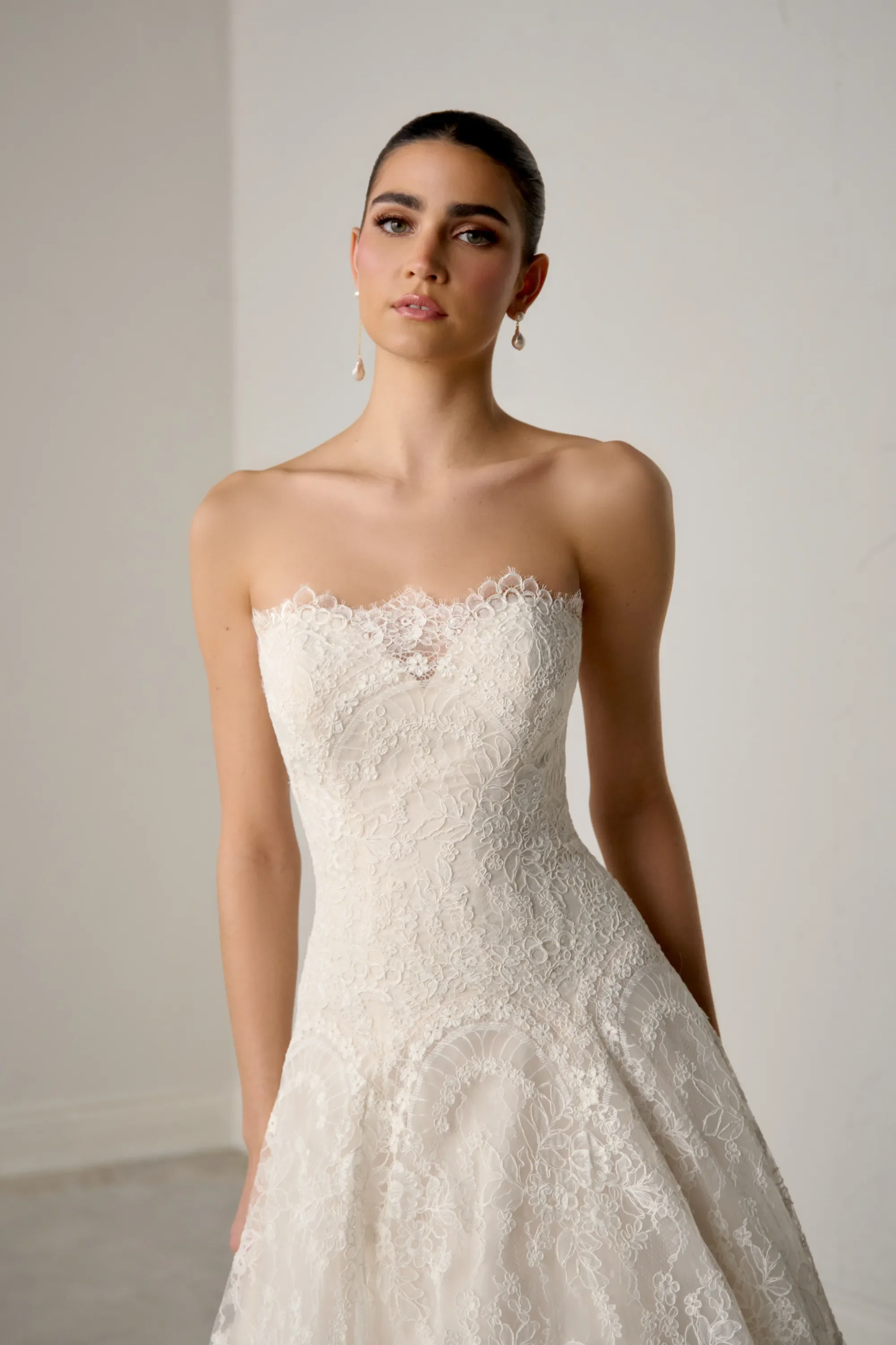 A young woman wearing a strapless white lace wedding gown with floral pattern, standing against a plain background.