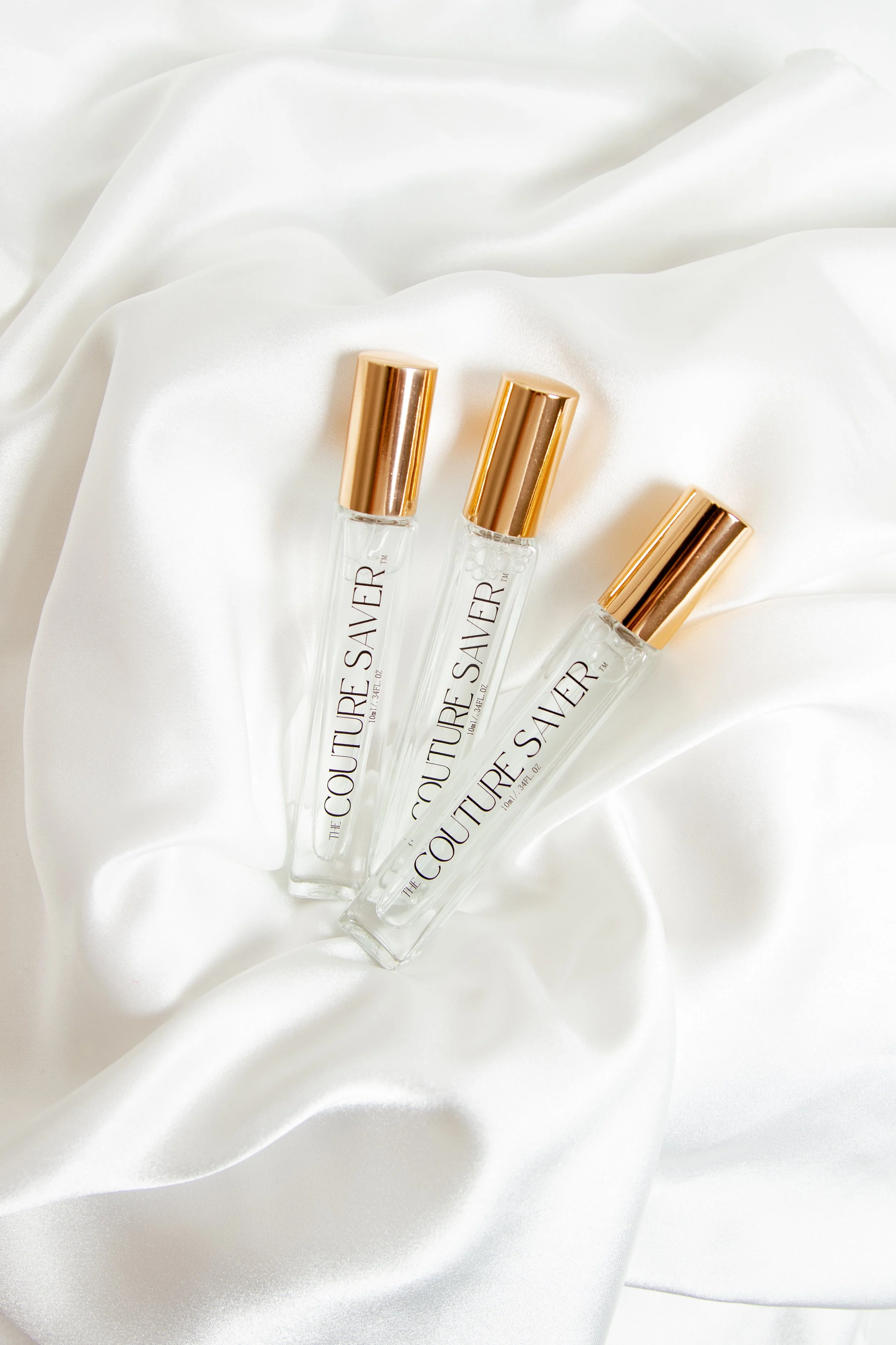 Three glass bottles of Couture Save perfume with gold caps on white satin fabric.