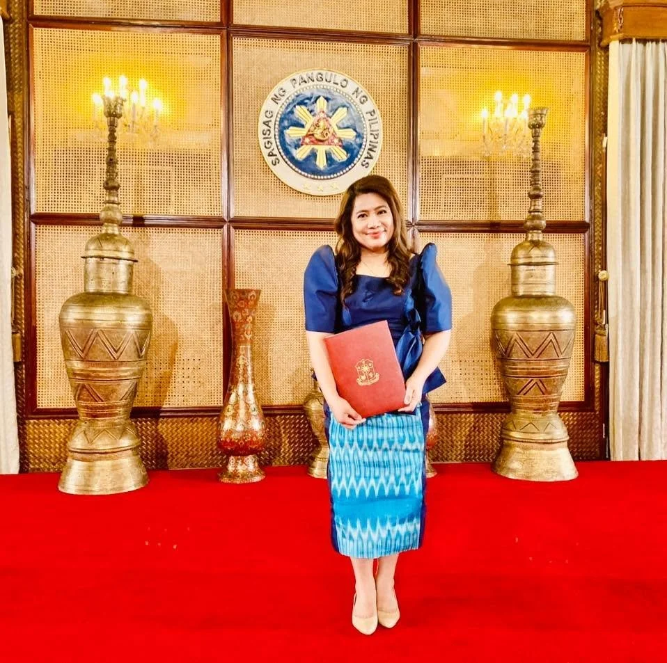 A woman wearing a blue traditional dress holding a red folder with a gold emblem, standing on a red carpet in front of a wooden wall with the Philippine seal, flanked by two large gold vases and candle holders.