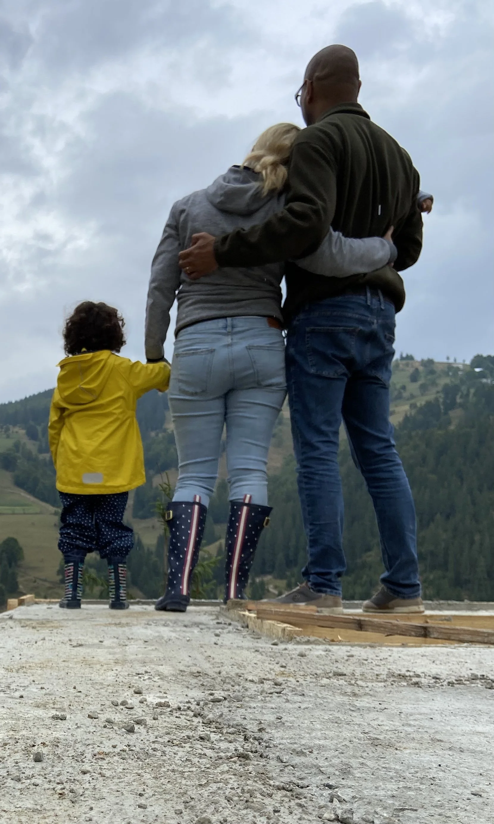 A family of three, including a child in a yellow raincoat, stand together outdoors on a cloudy day, facing away, overlooking a hilly landscape.