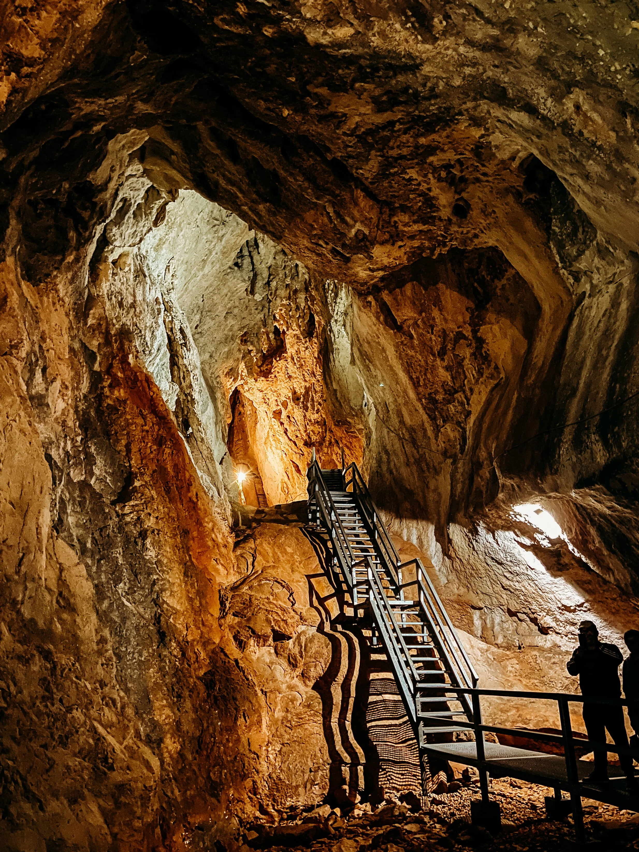 Inside a large, illuminated cave with orange and brown textured rock walls, a staircase leading upward, and a person standing at the bottom of the stairs.
