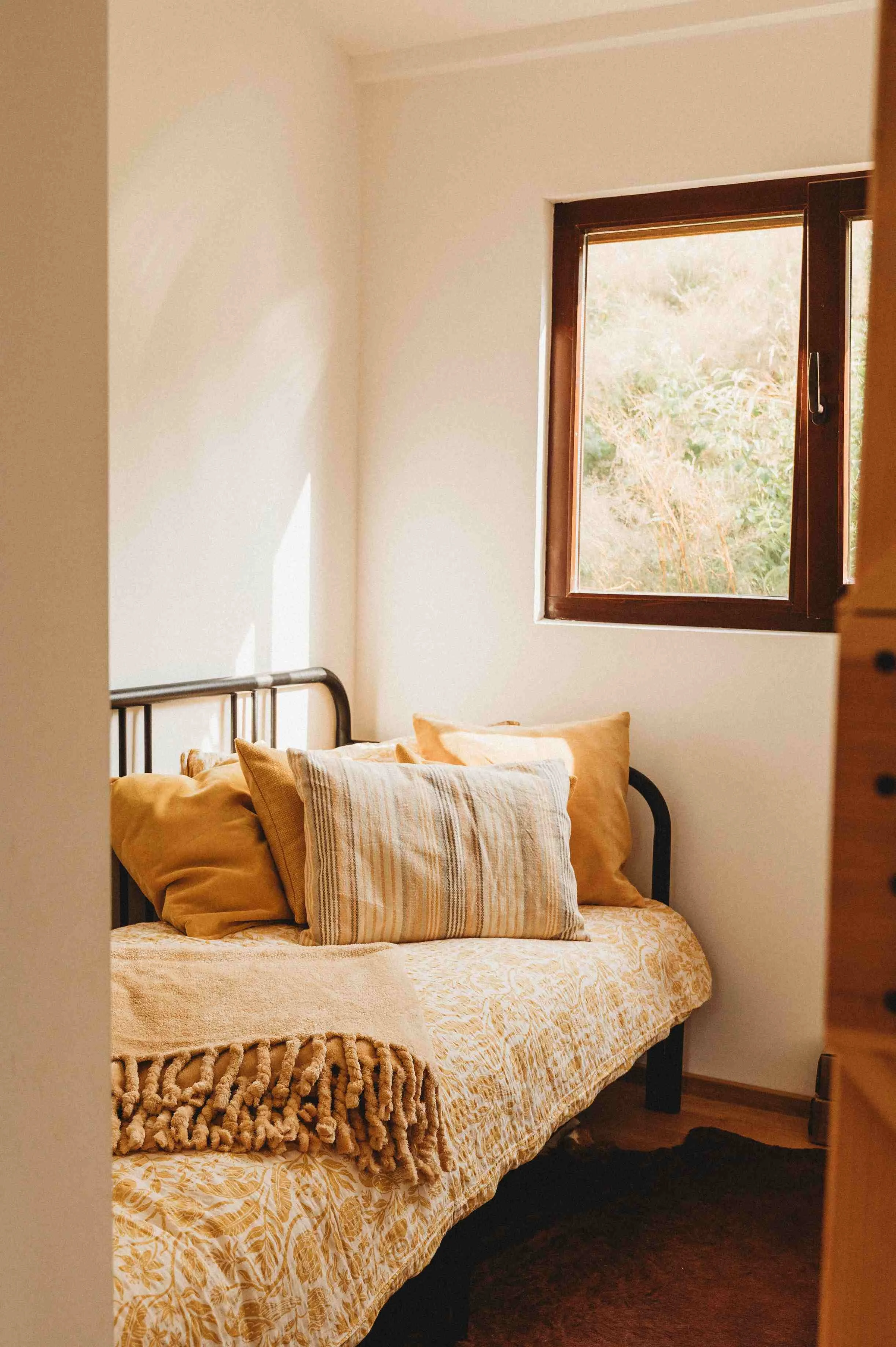 Cozy bedroom with a black metal bed frame, beige and striped pillows, a patterned bedspread, a window with brown trim showing greenery outside, and dark wood flooring.