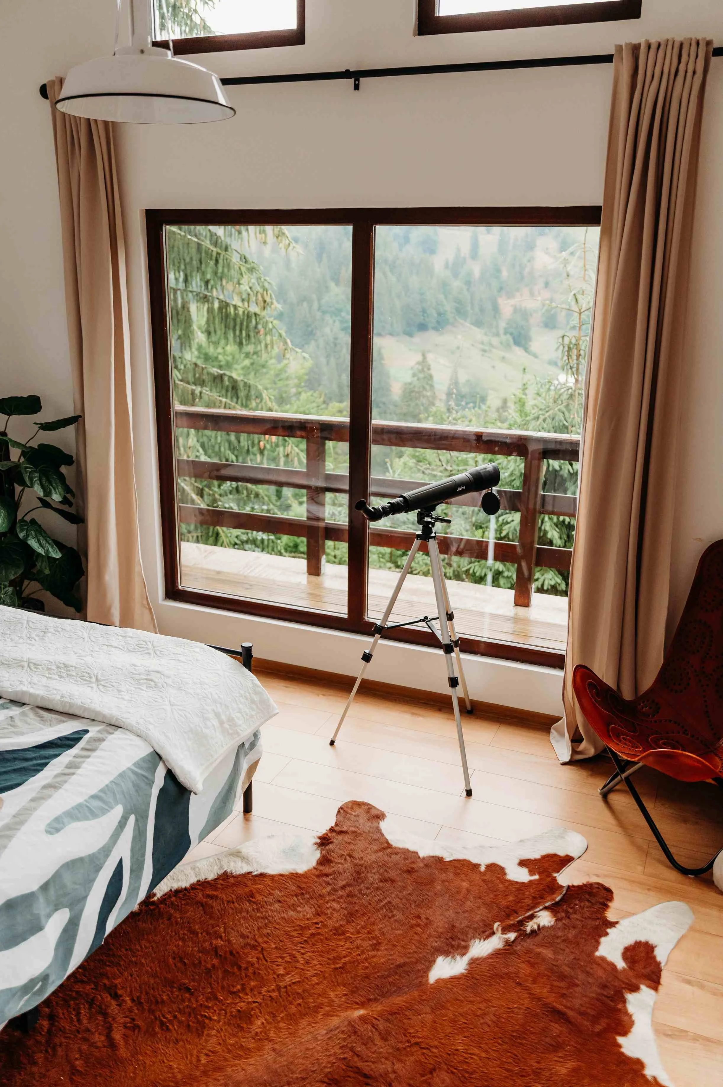 A bedroom with a large window and a sliding glass door opening to a balcony overlooking a green forest. There is a telescope on a tripod facing the window, beige curtains on both sides of the window, a bed with a white quilt and a blanket featuring a large leaf design, a green plant on the left, and a reddish-brown cowhide rug on the wooden floor.