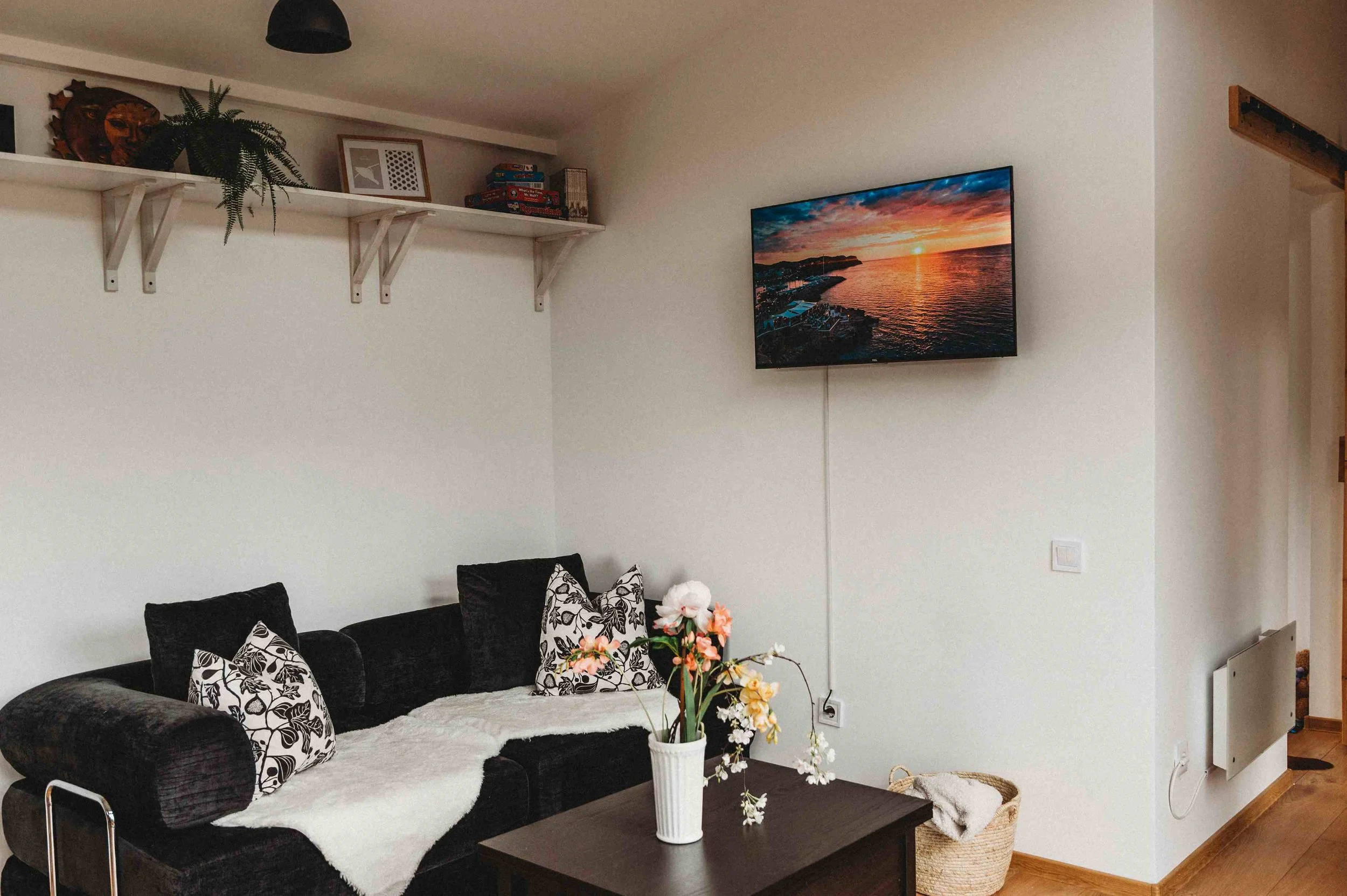 Living room with black sofa, decorative pillows, a white throw blanket, a coffee table with a potted plant, and a wall-mounted TV showing a sunset over water.