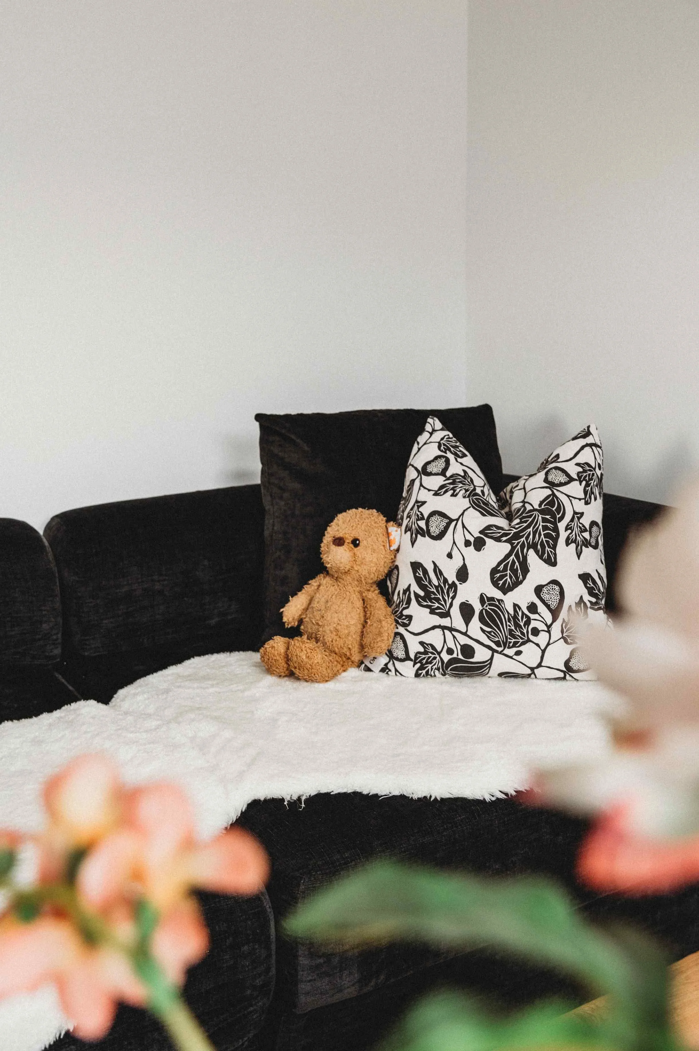 Close-up of decorative pillows on a dark sofa, with a plain wall in the background and a blurred floral element in the foreground.