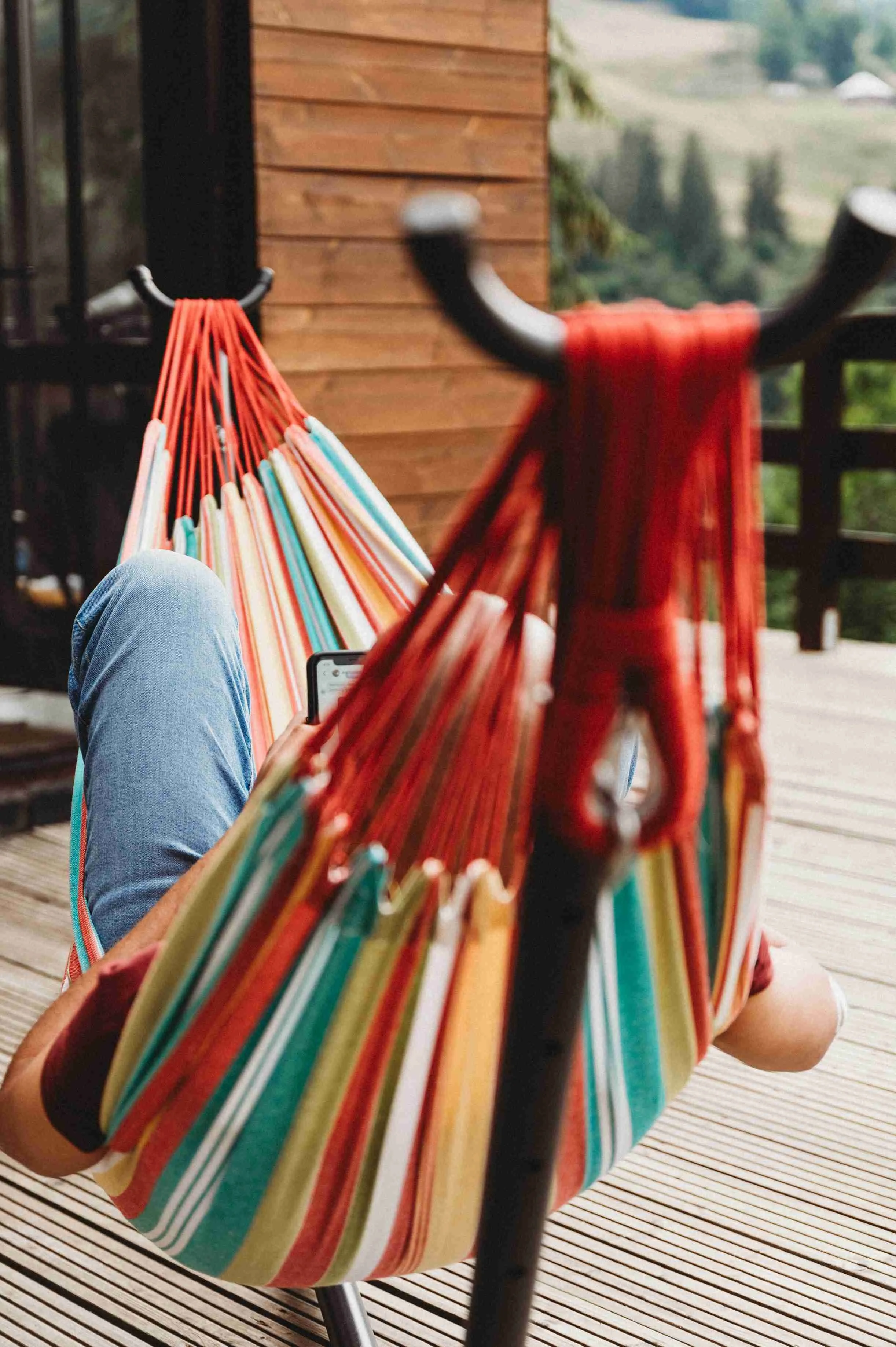 Person relaxing on a striped outdoor lounge chair wearing blue jeans, holding a smartphone, with a colorful hammock in the foreground, and a wooden wall with a scenic view of rolling green hills in the background.