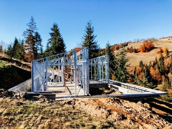 Construction site with metal framing for a building, surrounded by trees and hills under clear blue sky.
