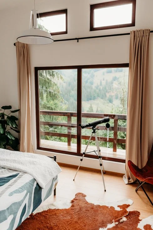 Bedroom with a large window overlooking a forested landscape, a telescope set up on a tripod, beige curtains, a bed with patterned bedding, a potted plant, a cowhide rug, and a wooden chair.