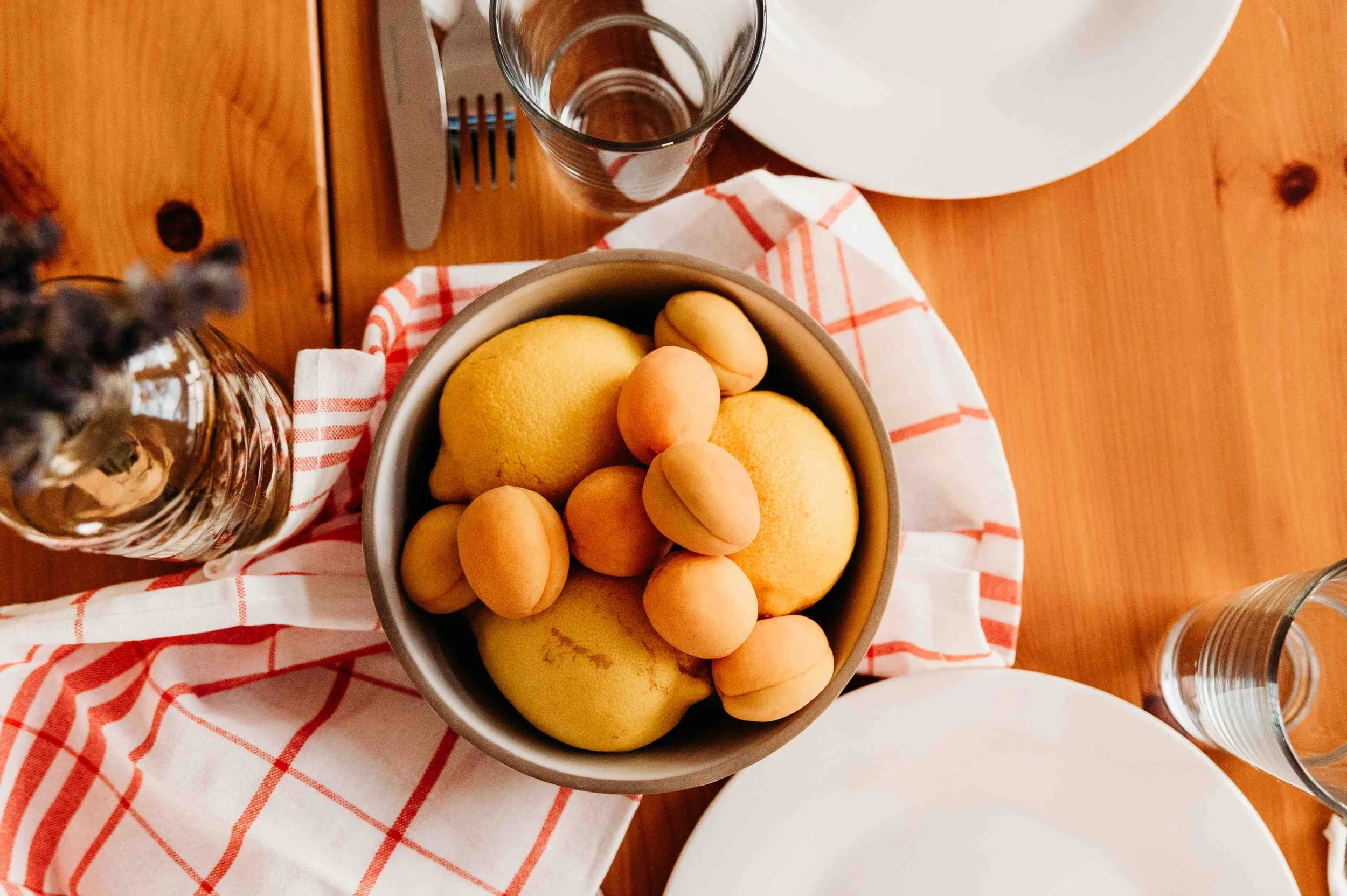 A bowl of assorted peaches and apricots on a table with a red and white checkered cloth, surrounded by empty glass cups and white plates.