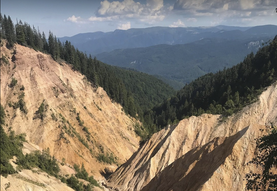 View of a mountain valley with layered, eroded rock formations in the foreground, dense forests on the slopes, and mountain ranges in the distance under a partly cloudy sky.