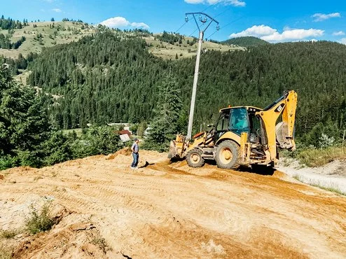 A construction worker standing next to a backhoe loader on a dirt road in a hilly, forested area with utility poles and blue sky background.