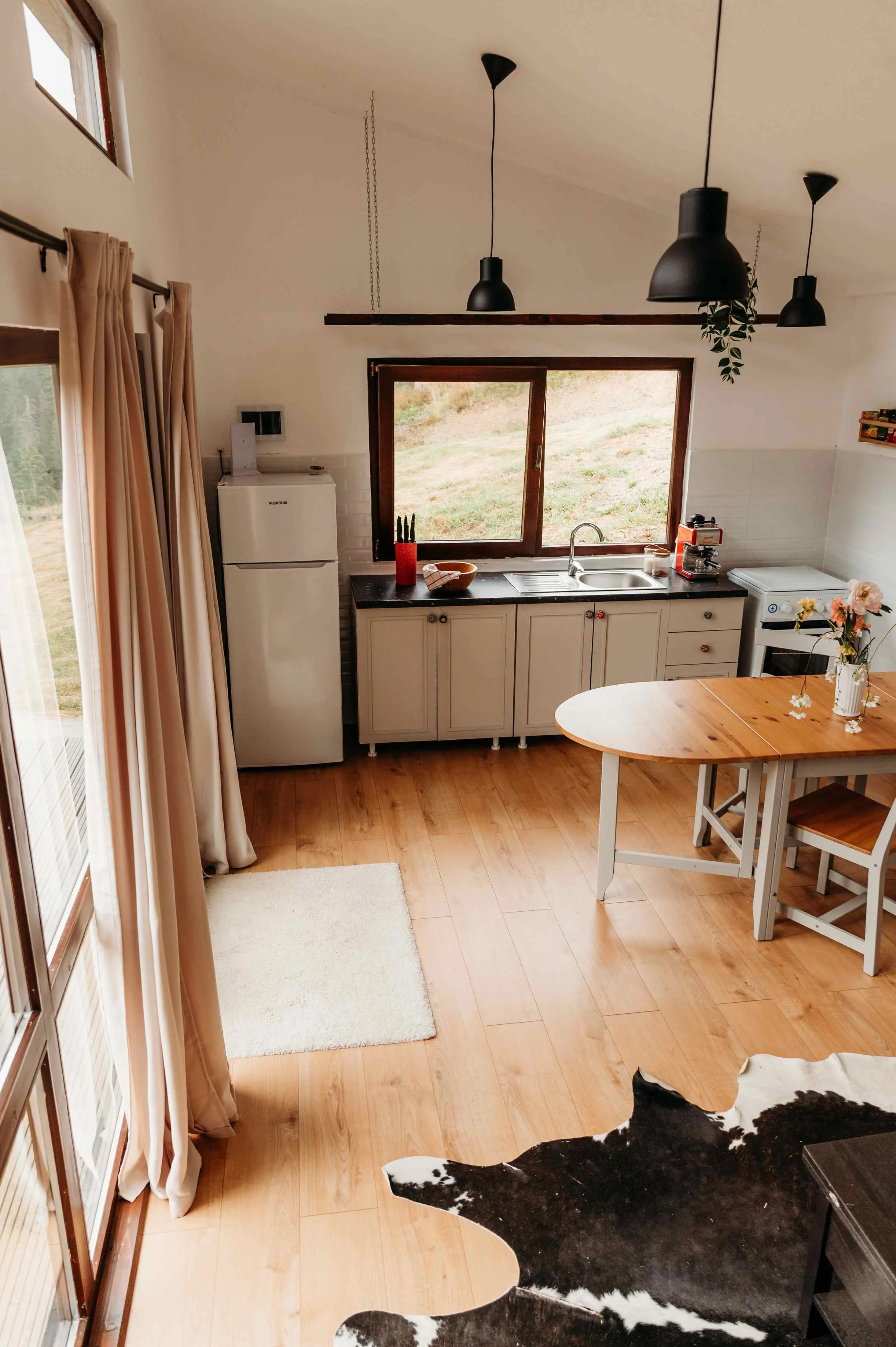 A kitchen with a window, beige curtains, a fridge, a black countertop with a knife holder, bowls, a coffee machine, and a vase with flowers. There is a round table with chairs, and part of a cowhide rug is visible on the wooden floor.