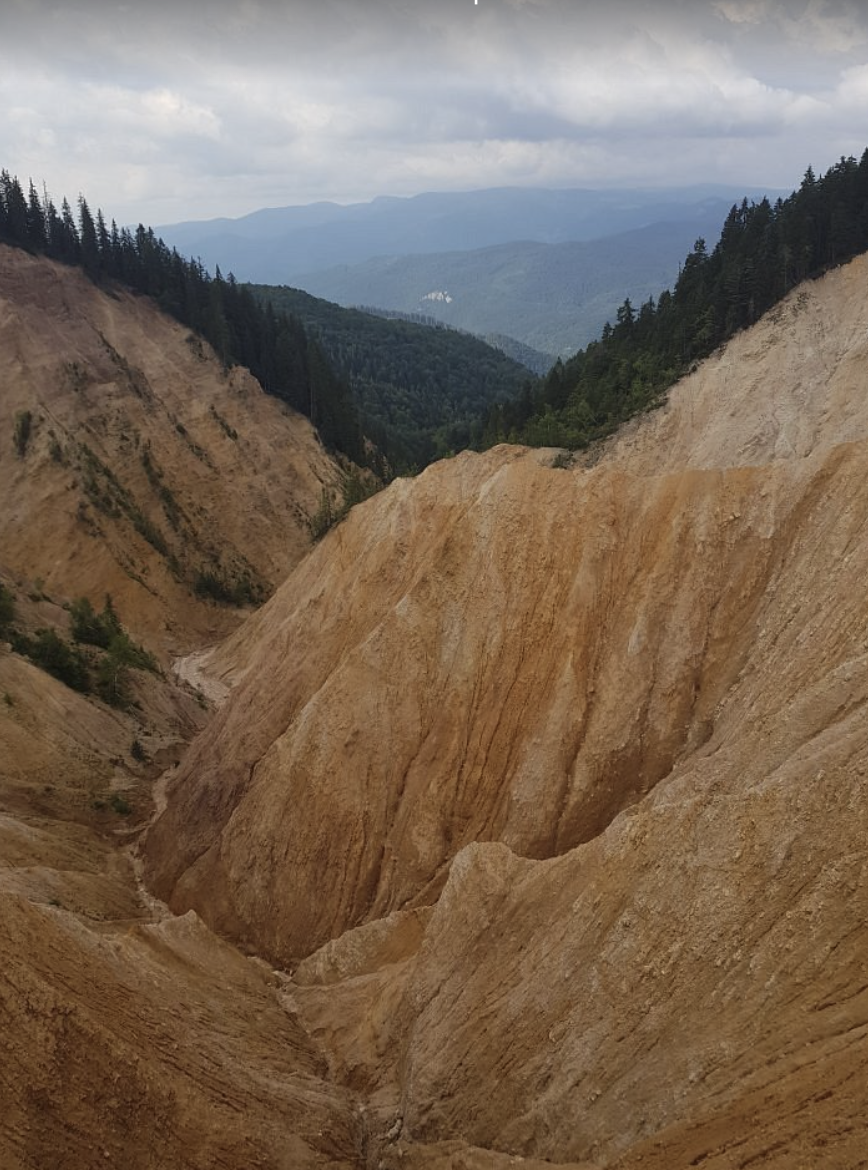 Photo of a deep canyon with rugged, orange-brown cliffs, dense green forested hills, and mountain ranges in the distance under a cloudy sky.