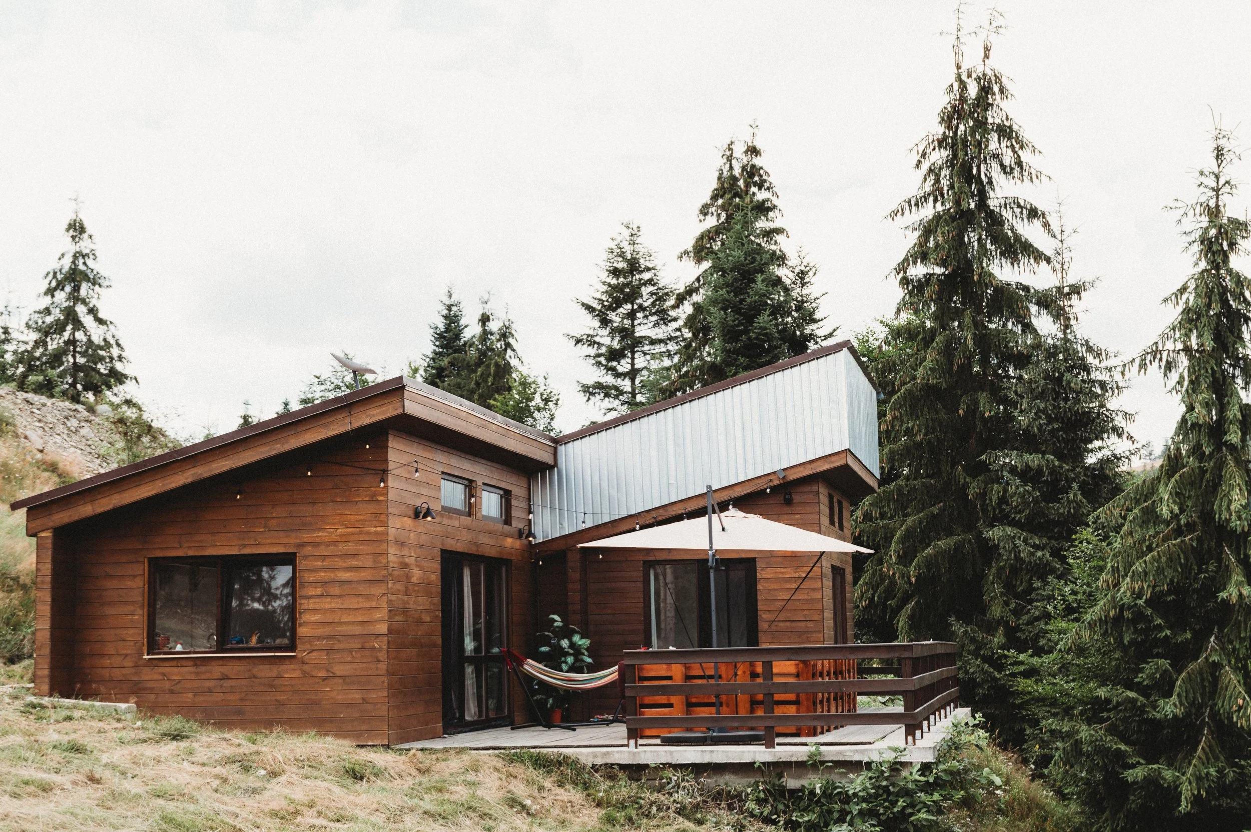 A modern wooden house with large windows, a deck with string lights, and a hammock on the porch, surrounded by tall pine trees under a cloudy sky.
