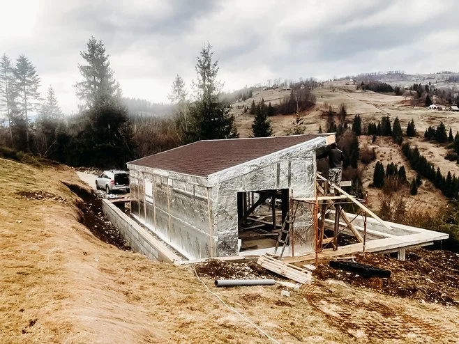 A house under construction on a hilly landscape with trees and a cloudy sky in the background.