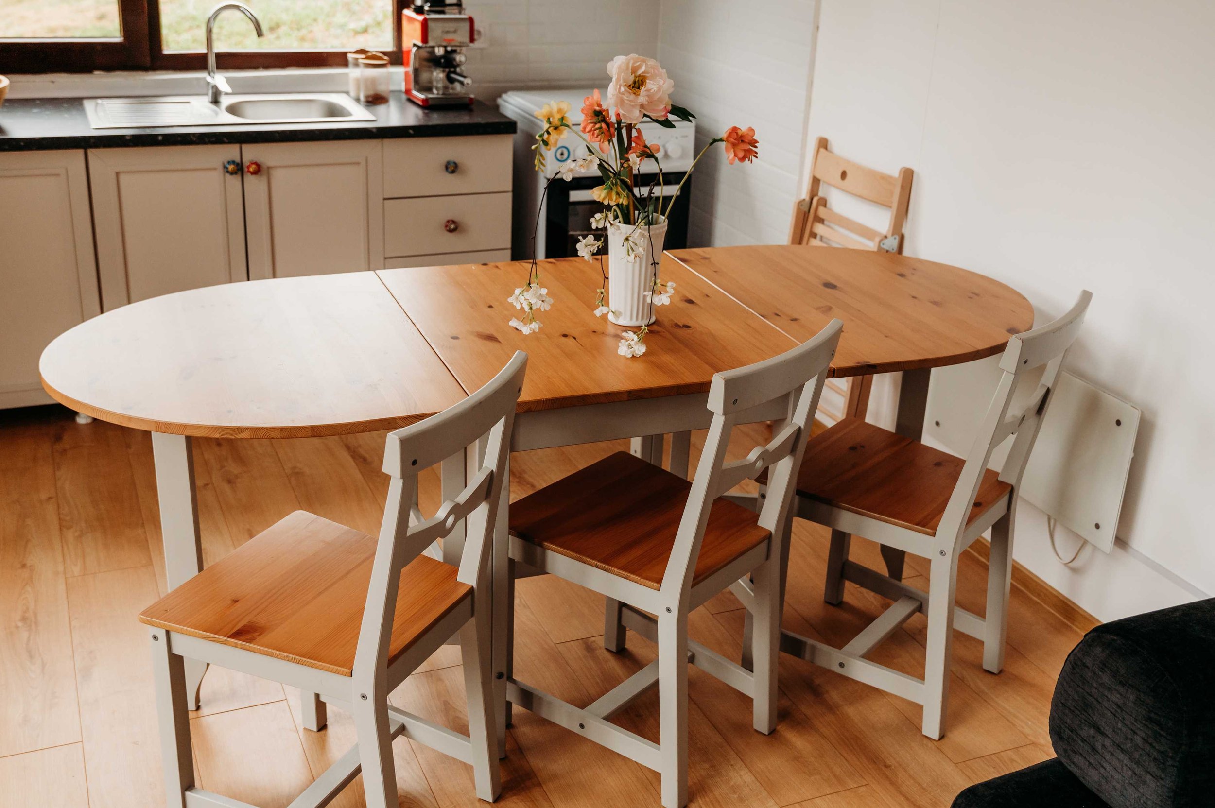 A wooden dining table with a flower vase with white and pink flowers in the center of the table, surrounded by white and brown chairs, in a kitchen with cream cabinets, a window, and some appliances.