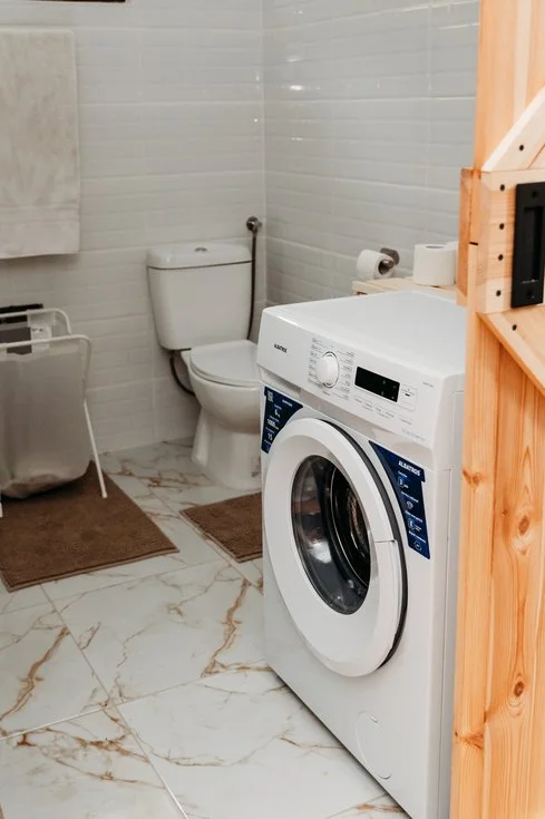 Bathroom with a toilet, washing machine, toilet paper, and brown rugs on marble floor.