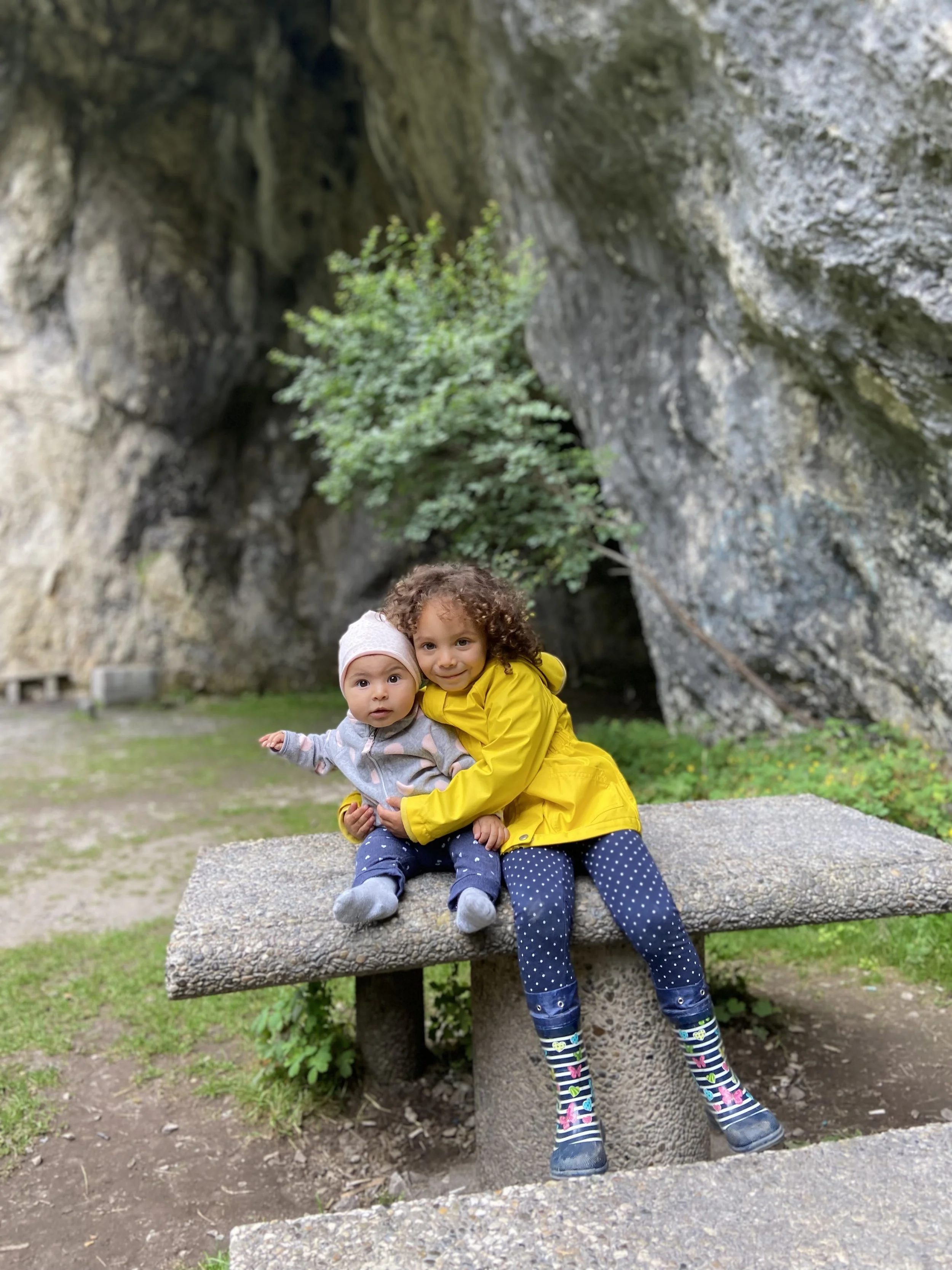 Two young children, a girl and a baby, sitting on a stone bench in front of a large rock formation and some greenery, with the girl holding the baby and both looking at the camera.