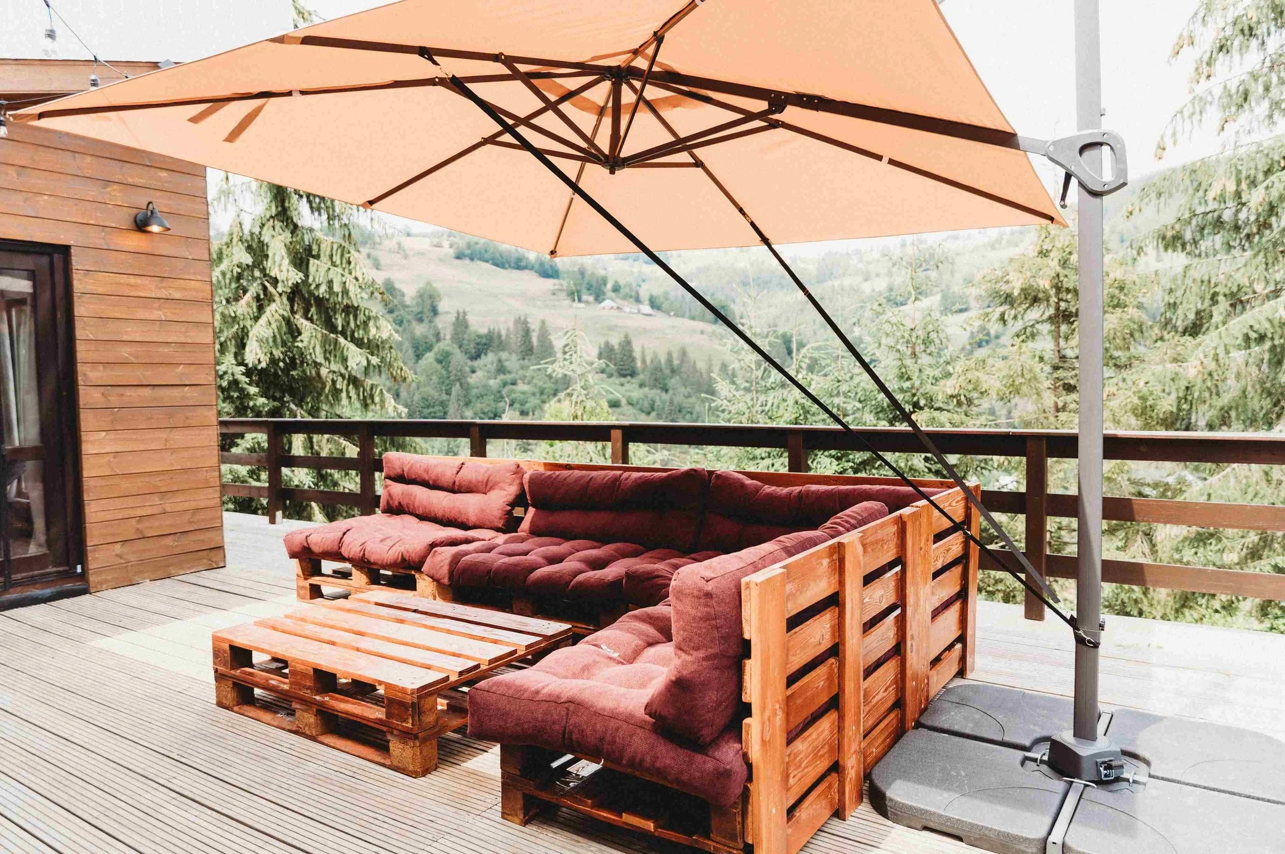 Outdoor patio area with a wooden sectional sofa with red cushions, a wooden coffee table, a large beige patio umbrella, a wooden house wall, and a scenic landscape of trees and hills in the background.