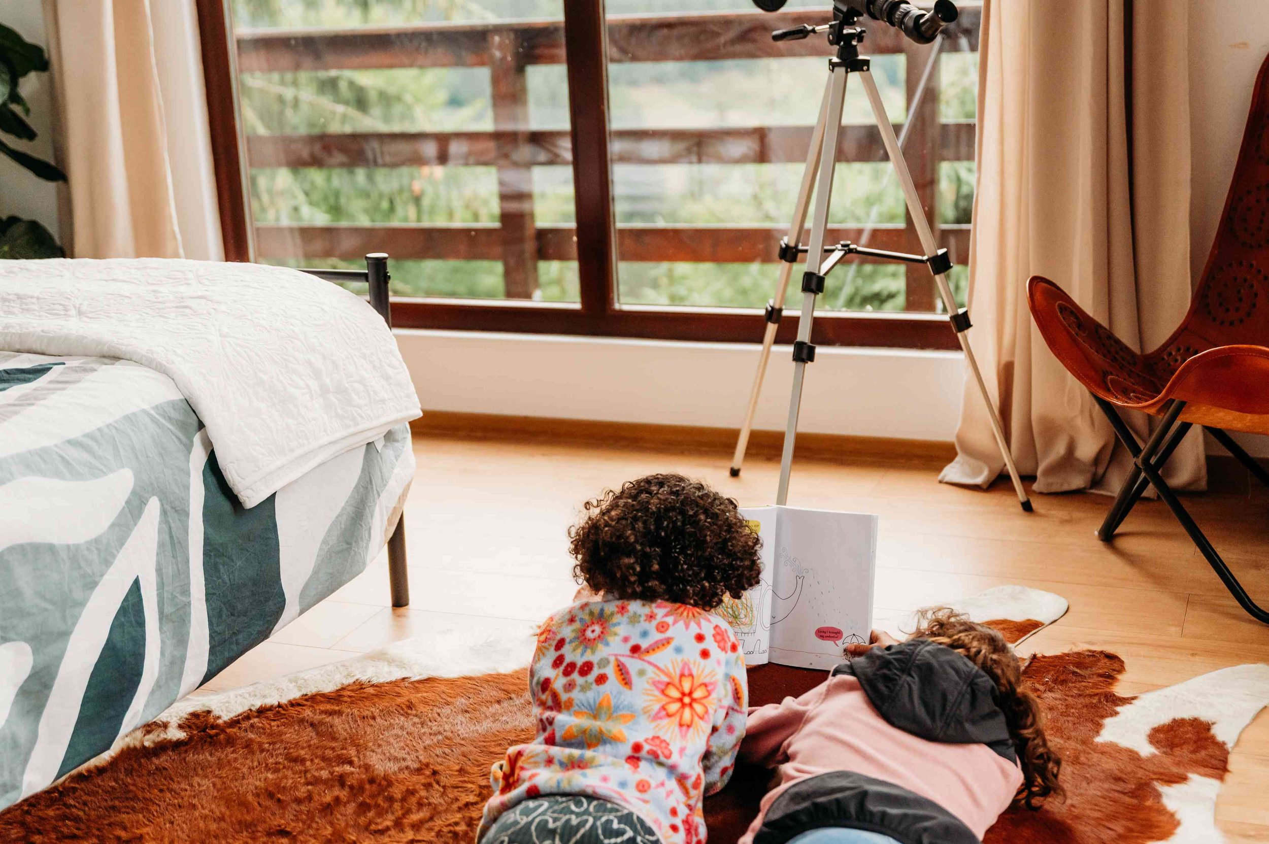 Two children lying on the floor looking at a book in a bedroom with large windows and a telescope.