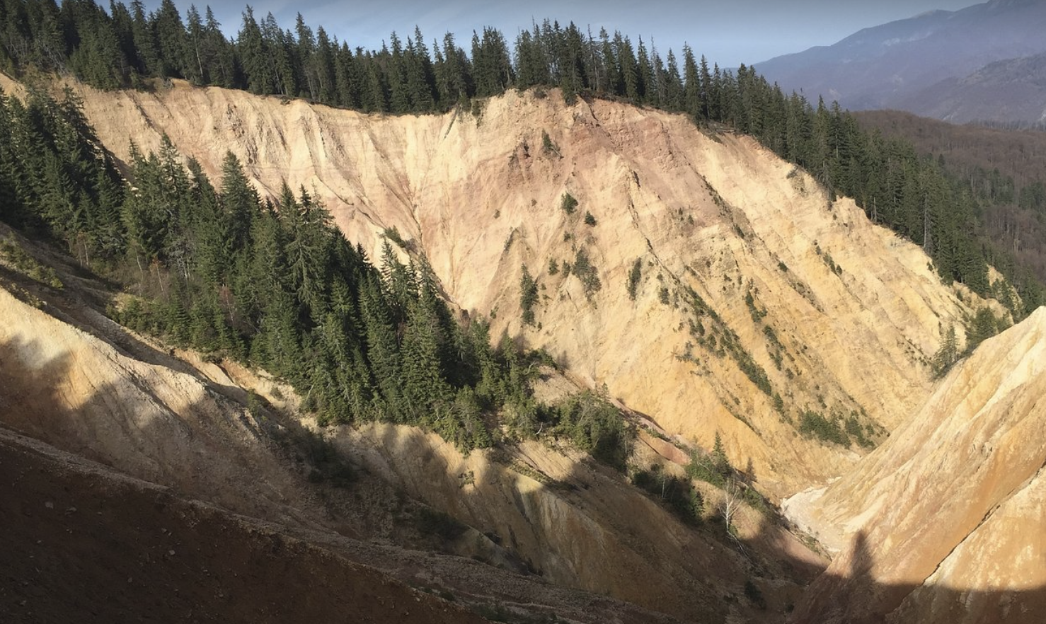 A rugged mountain landscape featuring steep, multicolored cliffs with patches of green pine trees, and distant mountains in the background.