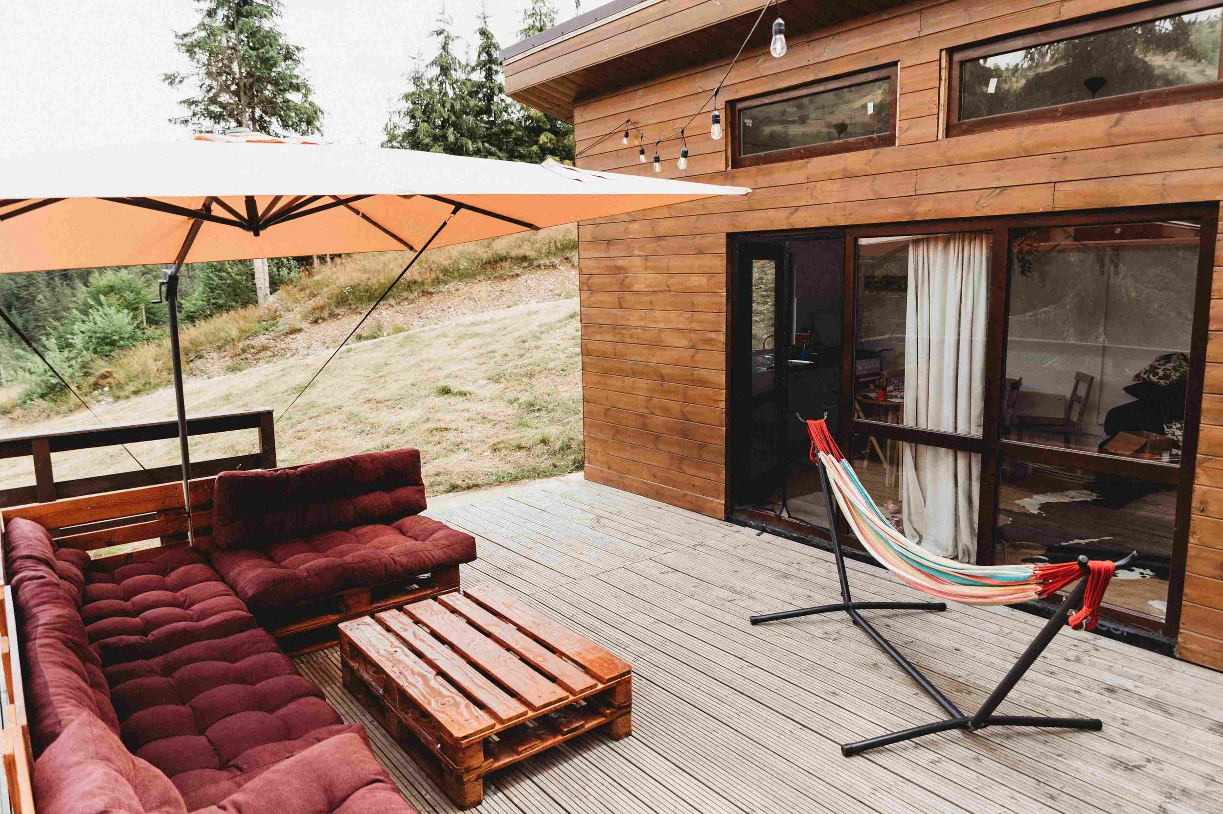 Outdoor wooden patio area with red cushioned seating, a wooden pallet coffee table, a beige patio umbrella, a hammock, and a string of lights in front of a modern wooden house with large windows and sliding glass door, surrounded by trees and greenery.