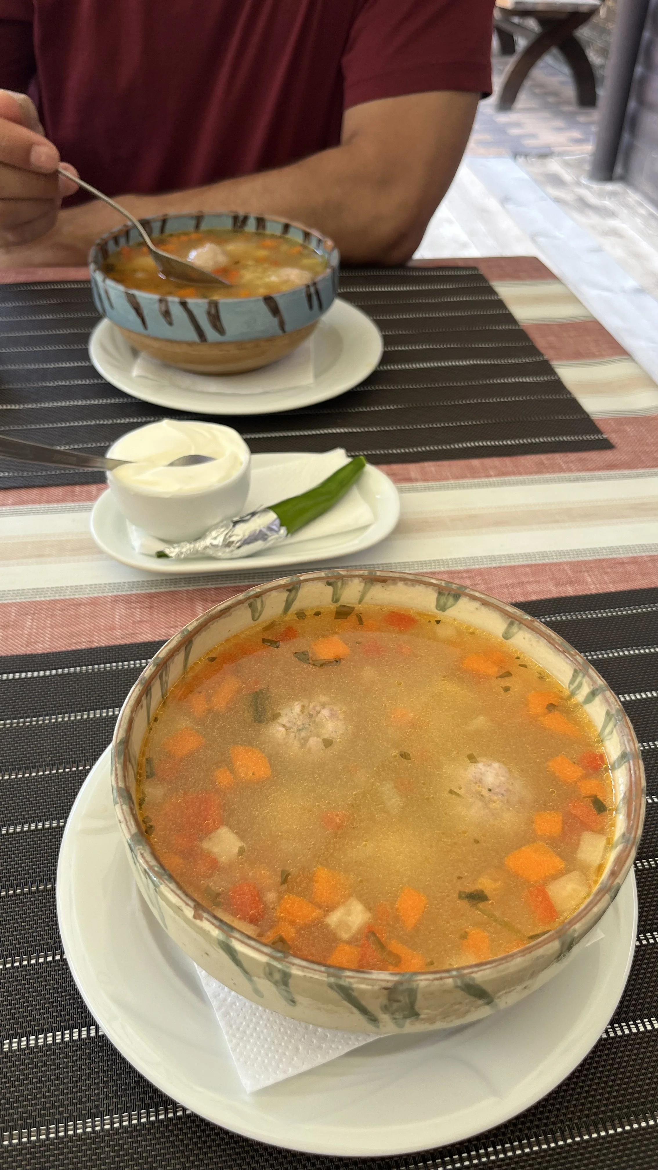 A bowl of vegetable soup with carrots, potatoes, and herbs on a white plate, with a spoon and a small dish of sour cream or yogurt with a green onion on a tablecloth. In the background, a person in a red shirt is eating and there is another bowl of soup.