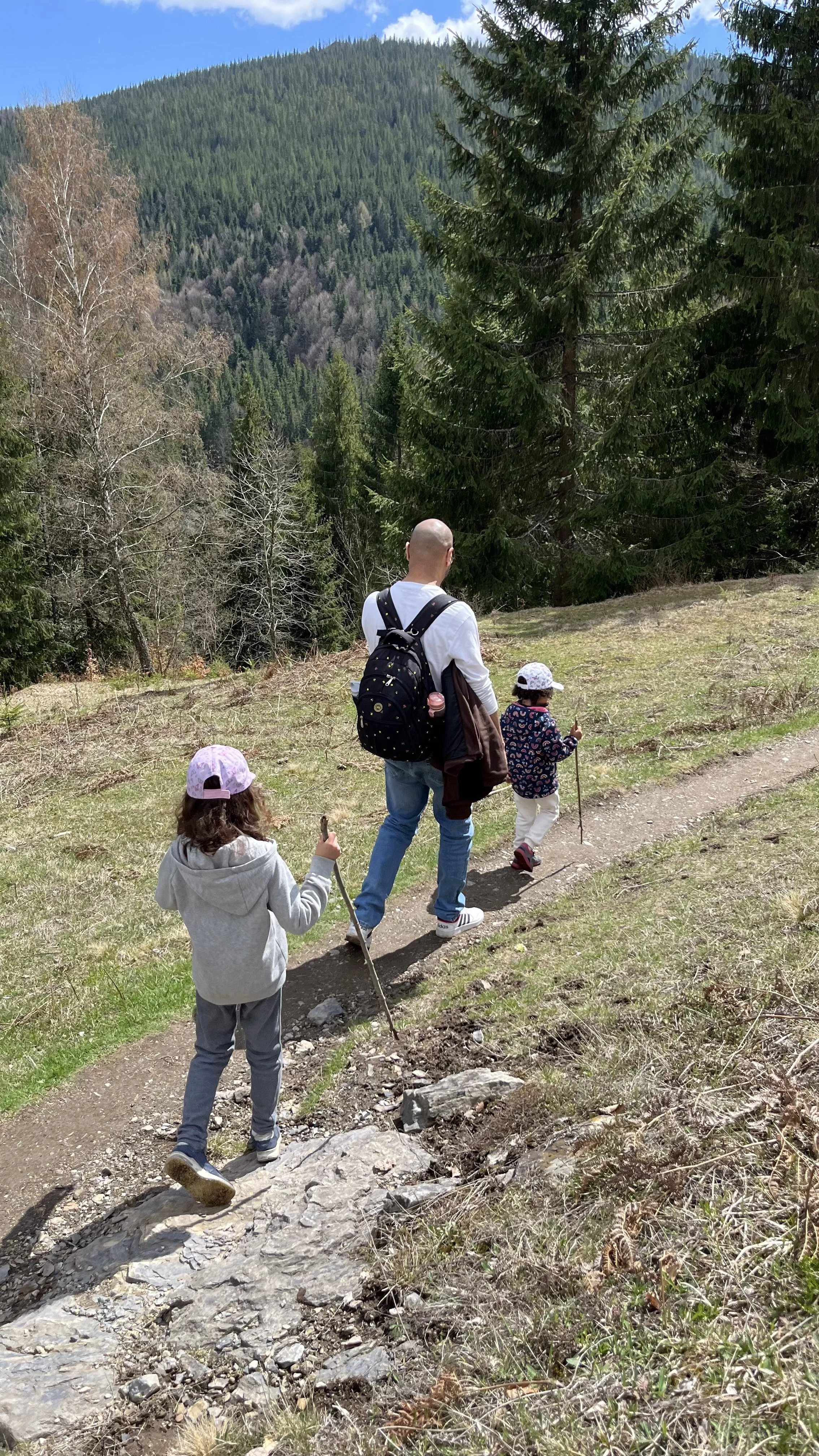 Bald man with a black backpack walking on a dirt trail with two young girls in a forested mountainous area on a sunny day.