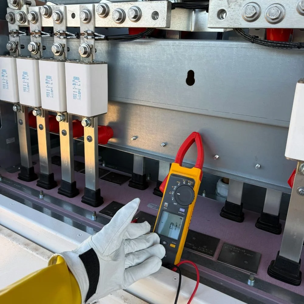 A person wearing a white glove and yellow sleeve holding a digital multimeter near electrical components inside an electrical panel or enclosure. The multimeter displays a reading of 0.1.