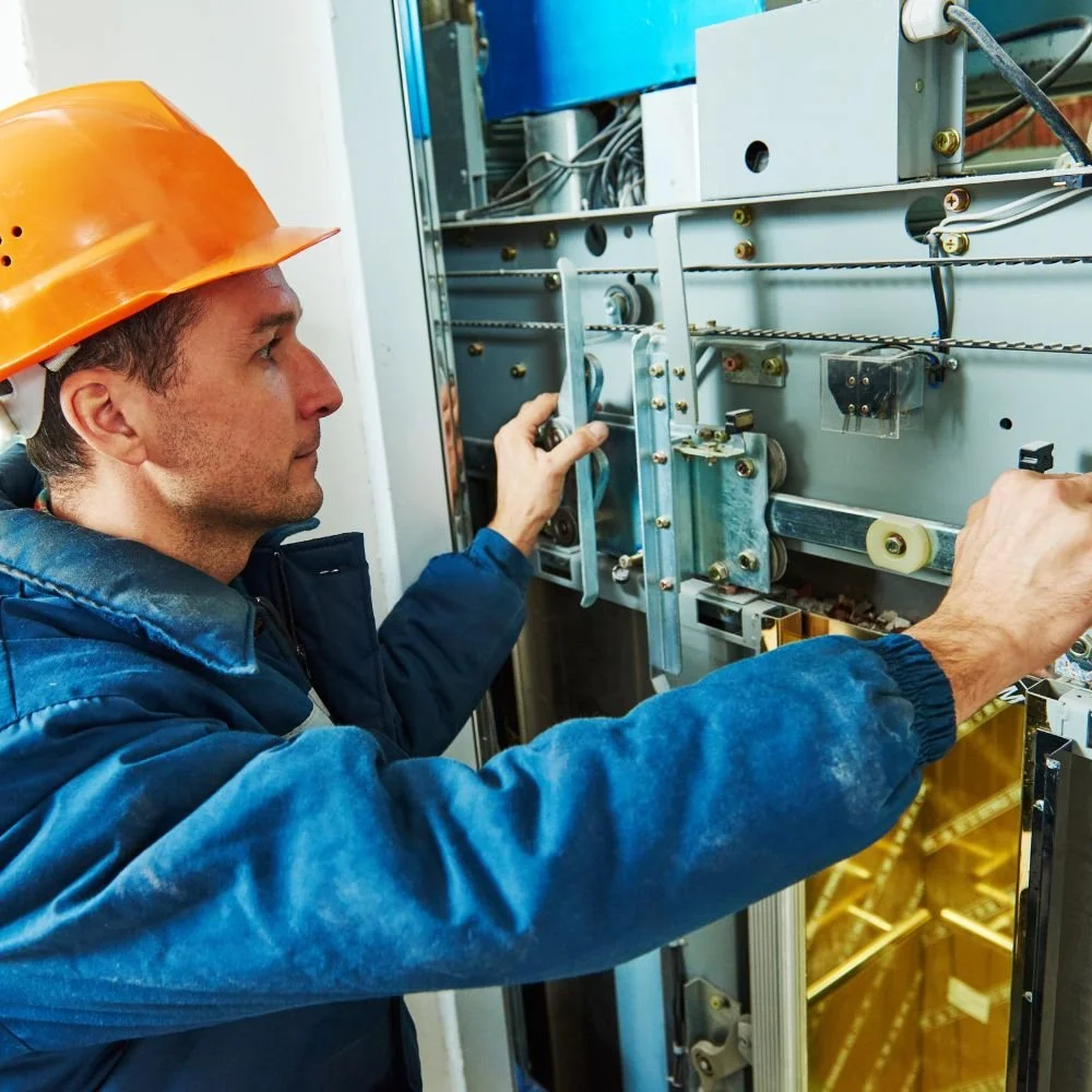 A man wearing an orange safety helmet and blue work jacket repairing or inspecting mechanical parts inside an industrial machine.