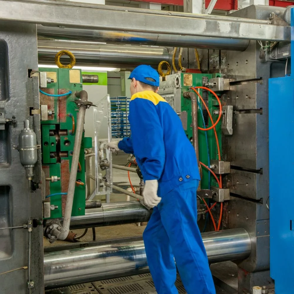 A worker in blue overalls and a blue cap operating a large industrial machine with metal and green components, surrounded by orange and blue cables.