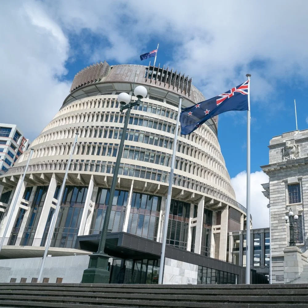 The Beehive, the New Zealand Parliament building, with two flags in the foreground, under a partly cloudy sky.