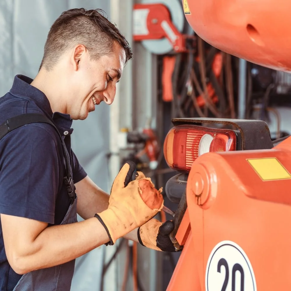 A young man wearing work gloves, smiling, as he works on an orange machine or vehicle with the number 20 on it, in an industrial or workshop setting.