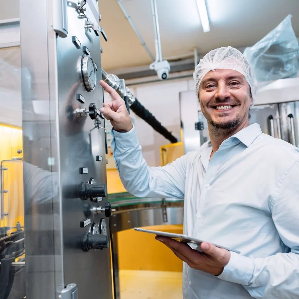 A smiling man in a white shirt and a hair net operating industrial machinery in a food or pharmaceutical manufacturing facility.